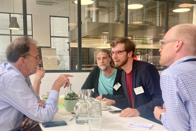 Eine Gruppe von Menschen sitzt um einen Tisch und führt eine Diskussion. Im Hintergrund ist ein modernes Küchenbereich mit schwarzen Fliesen zu sehen. Ein Mann mit Brille und Bart spricht zur Gruppe.